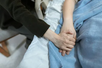 A caregiver sitting beside an elderly resident in a quiet, sunlit bedroom with a quilt on the bed and fresh flowers on the nightstand.