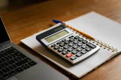A calculator, a notepad with handwritten cost figures, and a warm cup of coffee on a kitchen table.