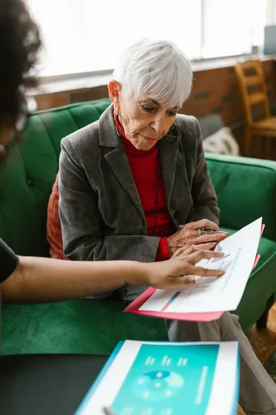 An older woman reading an insurance policy at a kitchen table with a pair of reading glasses and a cup of tea.
