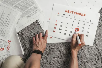 An older woman sitting at a kitchen table looking at a calendar with dates circled, a phone beside her.